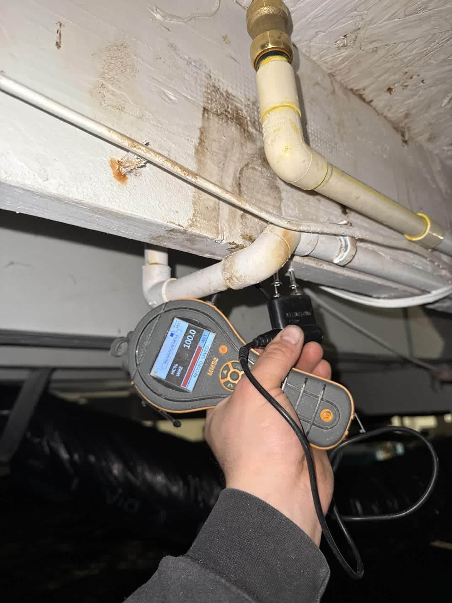 Moisture meter checking water-damaged framing near plumbing pipes.